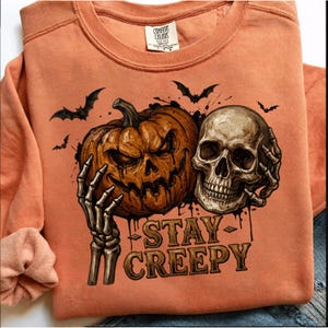 Peut inclure: Un sweat-shirt orange à col rond avec une impression graphique représentant une main de squelette tenant une citrouille sculptée et un crâne humain. De petites chauves-souris noires sont au-dessus. Le texte STAY CREEPY est imprimé en dessous.