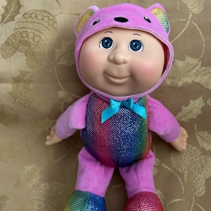 Può includere: Una bambola con un morbido cappuccio rosa a forma di orso con orecchie interne arcobaleno. La bambola ha occhi blu, carnagione chiara e un busto con motivo arcobaleno con un piccolo fiocco blu sul collo. I piedi sono coperti da tessuto luccicante.