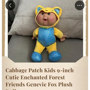 Könnte beinhalten: Eine 23 cm große Cabbage Patch Kids Cutie Enchanted Forest Friends Genevie Fox Plüschpuppe mit gelber Fuchskapuze, blauen Innenohren, blau glitzerndem Rumpf, gelben Gliedmaßen und blau glitzernden Füßen sowie einer kleinen grünen Schleife.
