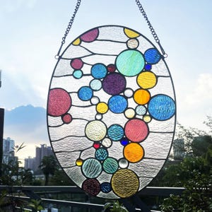 May include: An oval stained glass suncatcher featuring a composition of multicolored circular glass pieces in shades of blue, yellow, red, purple, and green, set against a clear textured glass background with a black metal frame and chain.