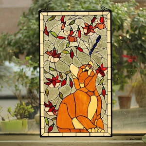 May include: A rectangular stained glass window hanging featuring an orange cat looking up at a blue dragonfly. The background consists of clear textured glass with red and white fuchsia flowers and green leaves, all framed by a black metal border.