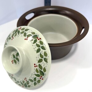May include: A round ceramic casserole dish with a dark brown exterior and a cream-colored interior. The matching lid features a cream base with a green leaf and red berry pattern, centered by a raised circular handle with a matching botanical design.