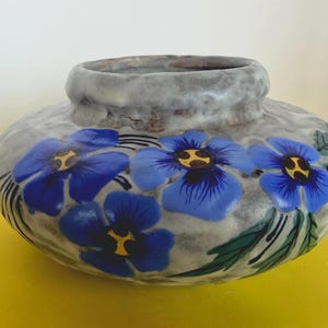 Puede incluir: Un jarrón de cerámica rechoncho y redondeado con un esmalte gris y blanco moteado. El exterior está decorado con flores azules pintadas a mano con centros amarillos y hojas de color verde oscuro. El jarrón tiene un borde ancho y grueso.