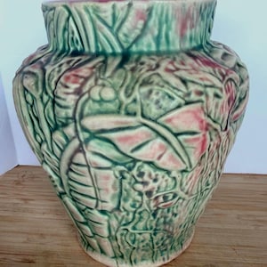 May include: A ceramic vase featuring a raised relief pattern of leaves and berries. The surface has a mottled glaze finish in shades of sage green and soft pink, with a wide opening and a tapered base that widens toward the middle of the body.