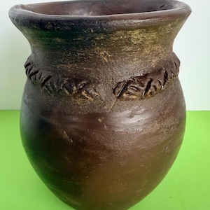 May include: A dark brown, rounded ceramic vessel with a wide, flared rim and a textured, braided-style band encircling the upper portion. The surface features a matte, earthy finish with subtle variations in tone and a visible, hand-crafted texture.