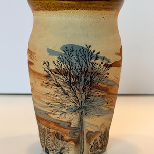 May include: A ceramic vase with a flared rim, featuring a marbled surface in shades of tan, brown, and light blue. The exterior is decorated with a detailed black silhouette of a large tree and smaller shrubs, set against the swirling clay colors.