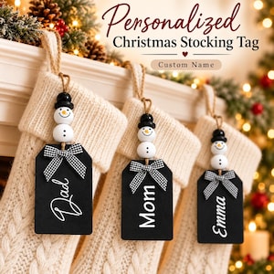 May include: Three black rectangular Christmas stocking tags with white text reading Dad, Mom, and Emma. Each tag features a snowman bead topper with a black hat and a black and white gingham ribbon bow, hanging from a cream-colored knit stocking.