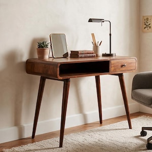 May include: A wooden desk with rounded edges, a single drawer, and four tapered legs. On the desk sits a small potted plant, a rectangular mirror, two stacked books, a pencil holder, and a black desk lamp. Beside it is a gray upholstered chair.