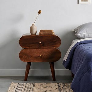 May include: A dark wood nightstand with two oval-shaped drawers and small gold knobs, positioned next to a bed with a blue quilted comforter and a grey pillow. A small beige vase with a dried plant sits on top of the nightstand.