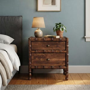 May include: A dark brown wooden nightstand with two drawers, each featuring a single round metal knob. The nightstand has a bamboo-style frame with rounded legs. A beige fabric lampshade sits on top, alongside a small potted green plant.