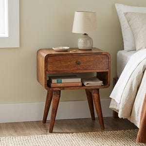 May include: A medium-brown wooden nightstand with rounded corners, featuring one drawer with a dark circular knob and an open storage shelf below. The nightstand stands on four tapered legs and holds a small lamp, a dish, and two books.