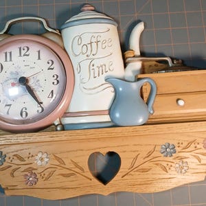 May include: A wooden wall shelf featuring a heart-shaped cutout and floral carvings. On the shelf sits a pink alarm clock, a white canister with the text Coffee Time, a small blue pitcher, and a wooden box with a drawer and a white knob.