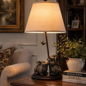 May include: A table lamp with a beige fabric shade and a dark bronze-toned base featuring two sculpted rabbits. The lamp has a thin metal stem, a round black base, and a pull chain with a small rabbit-shaped ornament hanging from the shade.