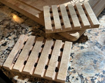 May include: Two rectangular soap dishes crafted from light-colored wood, featuring a slatted design with parallel horizontal bars and open gaps between them to allow for drainage. The surfaces show a natural wood grain texture.
