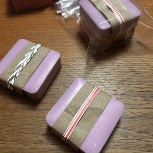 May include: Several square bars of lavender-colored soap wrapped in brown paper bands. Some bars are individually wrapped in clear plastic, while others are unwrapped. The paper bands feature thin red and white striped or leaf-patterned accents.