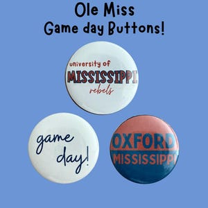 May include: Three round pinback buttons featuring the text Ole Miss Game day Buttons! at the top. One button is white with University of Mississippi Rebels text. Another is white with blue game day! text. The third is red and blue with Oxford Mississippi text.