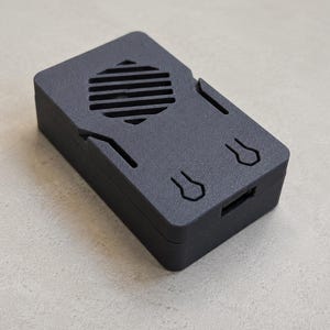 May include: A rectangular matte black protective case for a small electronic device. The top surface features a hexagonal ventilation grille with horizontal slats and two distinct keyhole-shaped cutouts, with a port opening on the side.