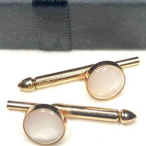May include: A pair of gold-toned cufflinks with a round, white, iridescent stone set in each cufflink. The cufflinks are displayed next to a black gift box with a black ribbon.