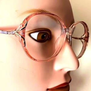 May include: Clear, oversized eyeglasses with a pink tint and decorative gold accents. The frames have a rounded shape and a patterned temple. The glasses are set against a neutral background.