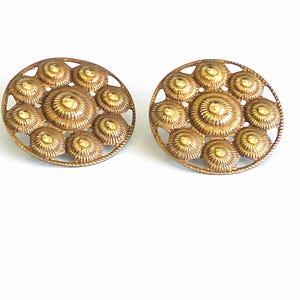 May include: A pair of circular gold-toned metal earrings featuring a central raised dome surrounded by a ring of eight smaller, textured, radiating circular motifs. The design includes an openwork border and a polished finish on the metal surface.