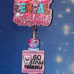 Op de afbeelding: Een badgehouder met een glitterende roze topper met de tekst DENTAL assistant, een afbeelding van tanden en een tube tandpasta. Daaronder bevinden zich kralen en een vierkante bedel met een lachende tandafbeelding en de tekst GO floss YOURSELF.
