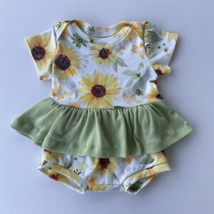 May include: A white baby onesie with a sunflower print and a green ruffle. The onesie has short sleeves and a ruffled bottom. The text "AMW handmade" is visible in the bottom right corner of the image.