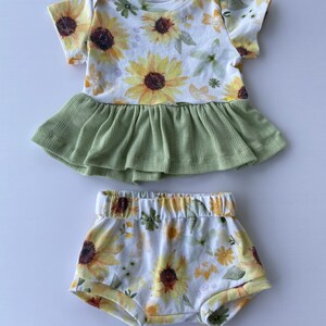May include: A baby outfit set with a sunflower print. The top is a short-sleeved bodysuit with a green ruffle skirt. The bottom is a pair of white shorts with a sunflower print.