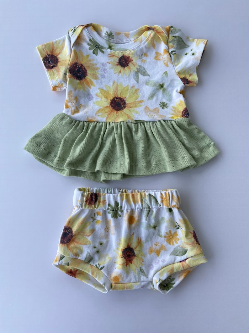 May include: A baby outfit set with a sunflower print. The top is a short-sleeved bodysuit with a green ruffle skirt. The bottom is a pair of white shorts with a sunflower print.