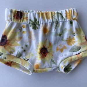 May include: White baby bloomers with a sunflower and floral print. The bloomers have an elastic waistband.