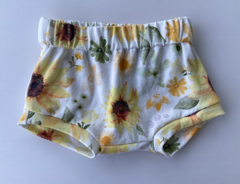 May include: White baby bloomers with a sunflower and floral print. The bloomers have an elastic waistband.
