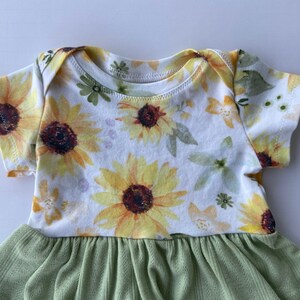 May include: A white baby onesie with a sunflower print and a green ruffled skirt. The onesie has short sleeves and a round neckline.