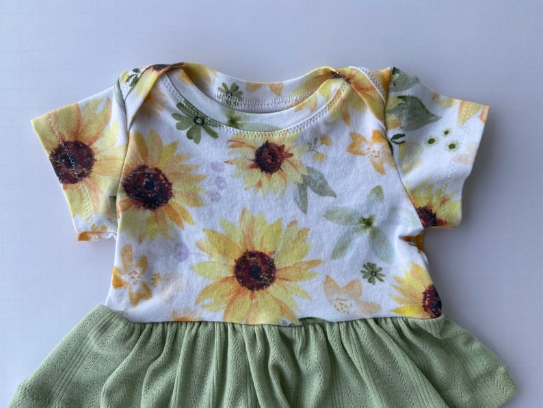 May include: A white baby onesie with a sunflower print and a green ruffled skirt. The onesie has short sleeves and a round neckline.
