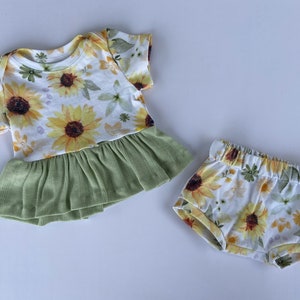 May include: A baby outfit set with a white short-sleeved shirt with a green ruffle and a white floral print with yellow sunflowers. The set also includes white shorts with a yellow sunflower floral print.