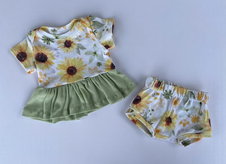 May include: A baby outfit set with a white short-sleeved shirt with a green ruffle and a white floral print with yellow sunflowers. The set also includes white shorts with a yellow sunflower floral print.