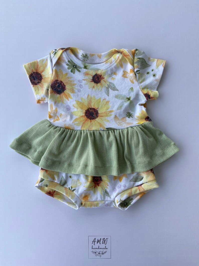 May include: A white baby onesie with a sunflower print and a green ruffle. The onesie has short sleeves and a ruffled bottom. The text "AMW handmade" is visible in the bottom right corner of the image.