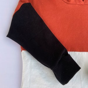 May include: A hooded sweatshirt with a colour block design. The sleeves are black, the top is orange, and the bottom is white.