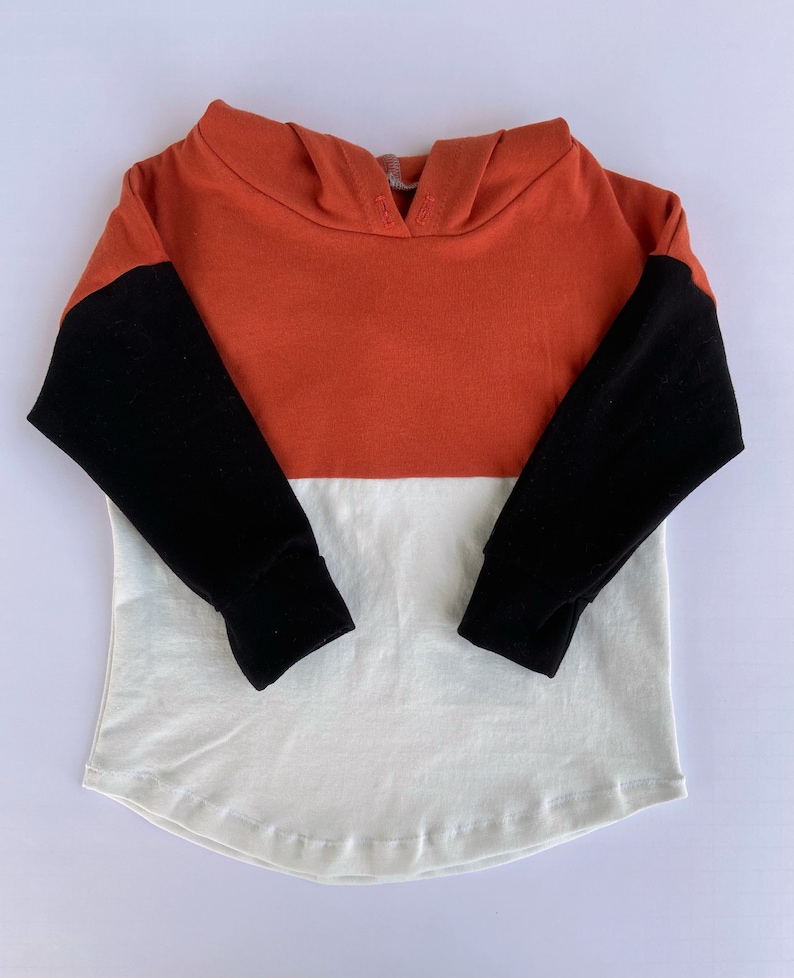 May include: A long-sleeved hooded sweatshirt with a colour block design. The sleeves are black, the top portion is rust orange, and the bottom portion is white.