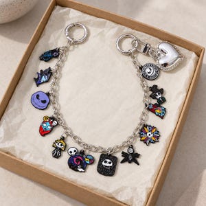 May include: A silver-toned metal charm bracelet featuring various colorful enamel pendants inspired by The Nightmare Before Christmas, including character faces, a heart-shaped clasp, and circular text charms, all arranged on a sturdy link chain.