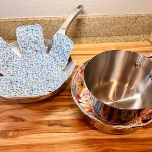 Puede incluir: Dos sartenes de acero inoxidable con protectores de tela. Un protector presenta un patrón floral azul y blanco, mientras que el otro tiene un estampado floral multicolor. Los protectores se colocan dentro de las sartenes para evitar rayones.