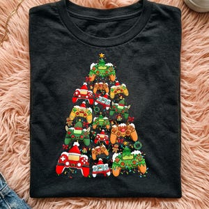 May include: A black short-sleeved t-shirt featuring a graphic print of a Christmas tree constructed from various colourful video game controllers. The controllers are decorated with Santa hats, reindeer antlers, and festive holiday lights.