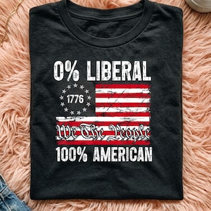 May include: A black t-shirt featuring a distressed graphic of the American flag with the text 0% LIBERAL at the top, 1776 inside a circle of stars, We The People in script, and 100% AMERICAN at the bottom in a bold, white, distressed font.