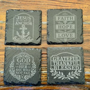 Op de afbeelding: Vier vierkante onderzetters van zwarte leisteen met ruwe, natuurlijke randen. Elke onderzetter is voorzien van lasergravures: Jesus Is My Anchor met een anker, Faith Hope Love met een kruis, God Is Within Her, en Grateful Thankful Blessed.