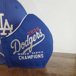 May include: A blue face mask with the text "Dodgers 2020 World Series Champions" in white and red. A blue baseball cap with a white "LA" logo is partially visible in the background. The mask is made of fabric.