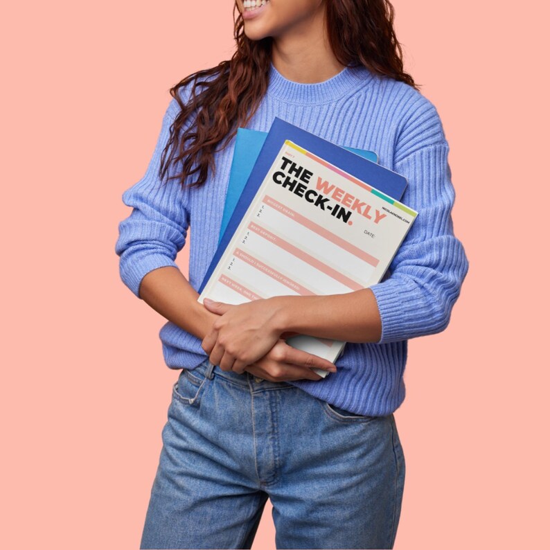 May include: A light blue ribbed sweater and blue jeans. The sweater is worn over a blue folder and a white planner with the text THE WEEKLY CHECK-IN printed in black and pink. The planner features horizontal pink lines for writing tasks.
