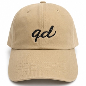 May include: A tan baseball cap featuring the letters qd embroidered in black script on the front center. The cap has a curved brim, a structured crown with eyelets, and a button at the top. The fabric appears to be a cotton twill material.