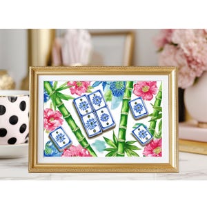 May include: A framed print featuring a watercolor design of blue and white dominoes, pink flowers, green bamboo stalks, and blue floral accents. The print is set in a gold frame and rests on a white surface.