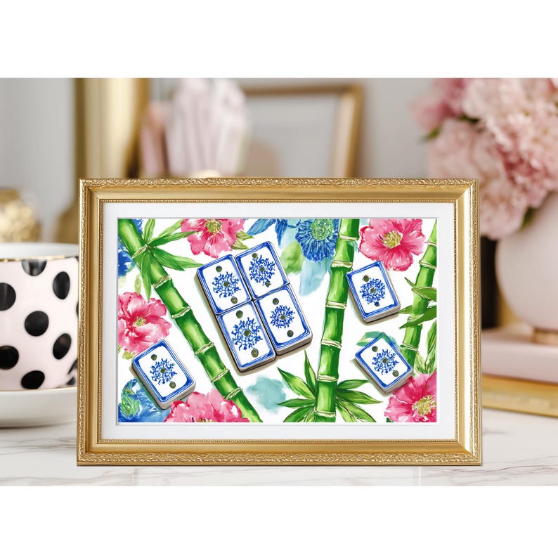 May include: A framed print featuring a watercolor design of blue and white dominoes, pink flowers, green bamboo stalks, and blue floral accents. The print is set in a gold frame and rests on a white surface.