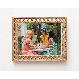 May include: A framed print depicts four women gathered around a table, playing a game. The women are dressed in colorful dresses. The frame is made of woven, light brown material. The background shows palm trees and a bright sky.