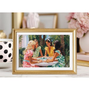 May include: A framed print depicts four women gathered around a table, playing a game. The artwork features a gold frame and a colorful scene with palm trees in the background. The women are dressed in various pastel-colored dresses.