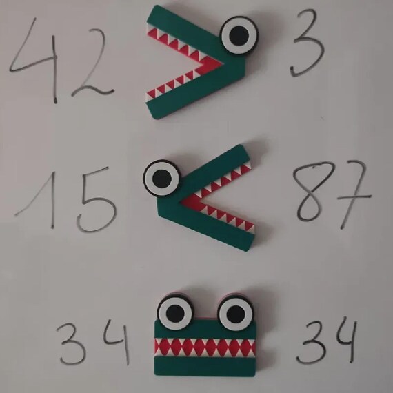 Three ChompCompare crocodile symbols on a whiteboard demonstrating correct greater-than, less-than, and equal number comparisons.